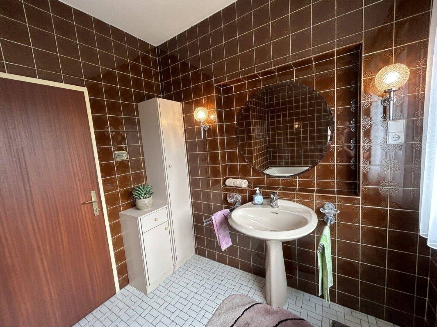 Badezimmer mit braunen Fliesen, einem Waschtisch mit Spiegelschrank und Wandleuchten.