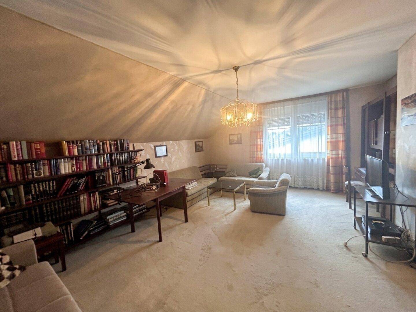 Helles Wohnzimmer im Dachgeschoss mit Schrägen, Teppichboden und einem großen Fenster.