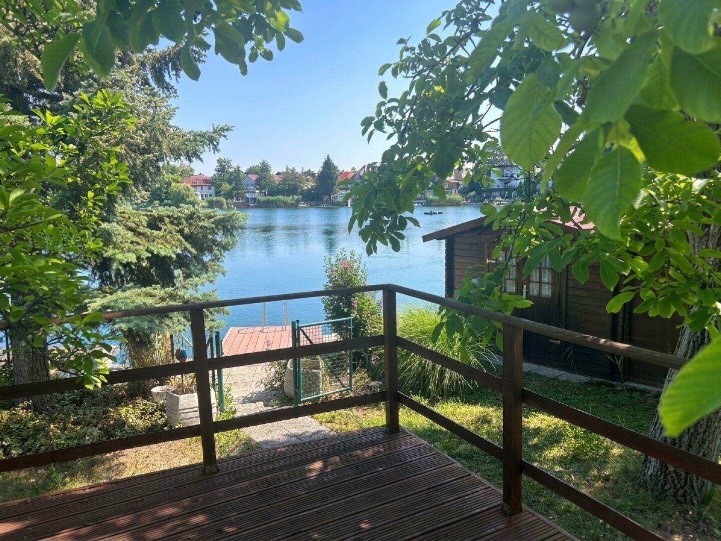 Holzterrasse mit Blick auf den See, umgeben von üppigem Grün und einem kleinen Holzhaus.