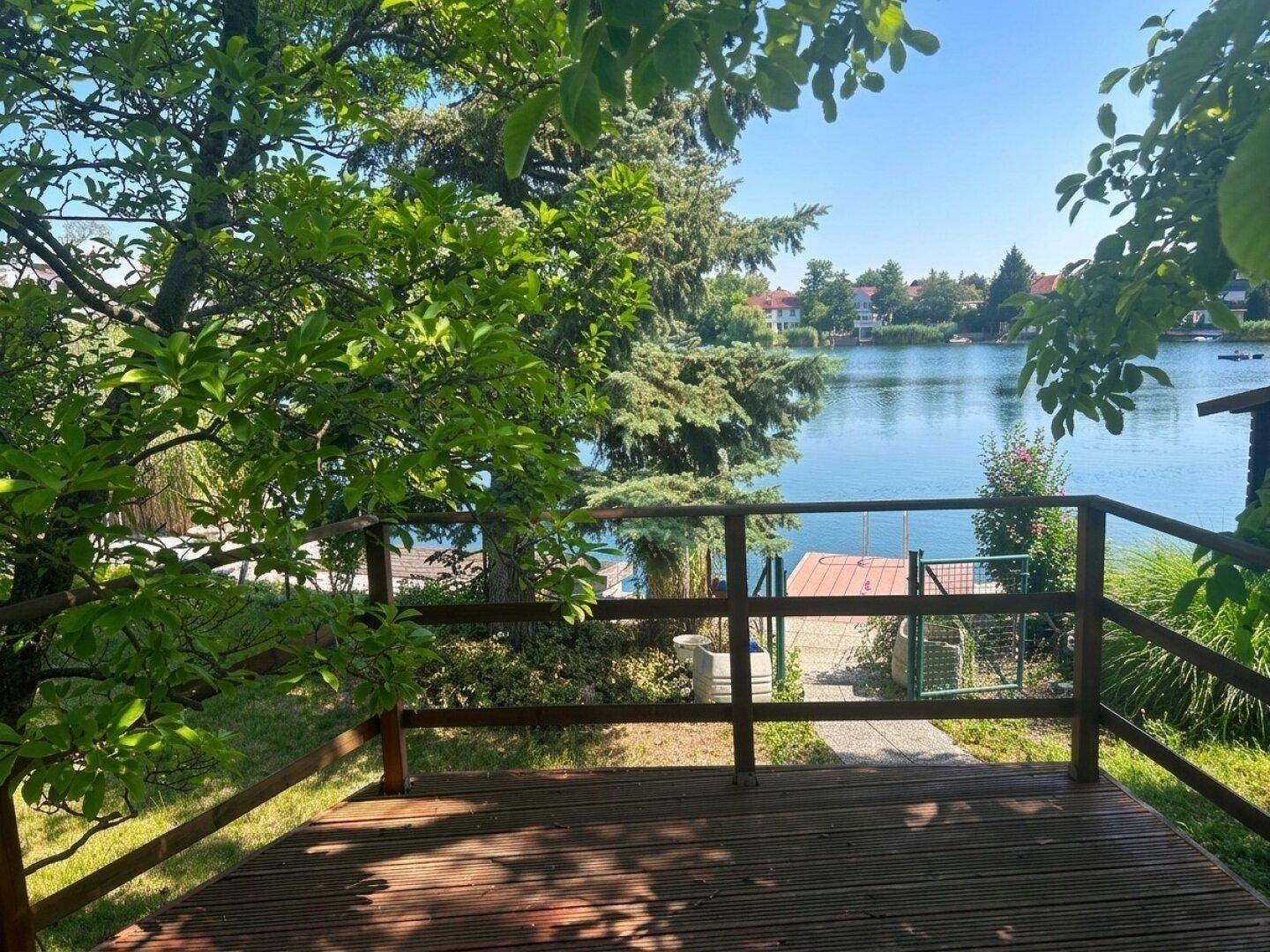 Holzterrasse mit Zugang zum See und einem kleinen Steg, umgeben von Bäumen und Pflanzen.