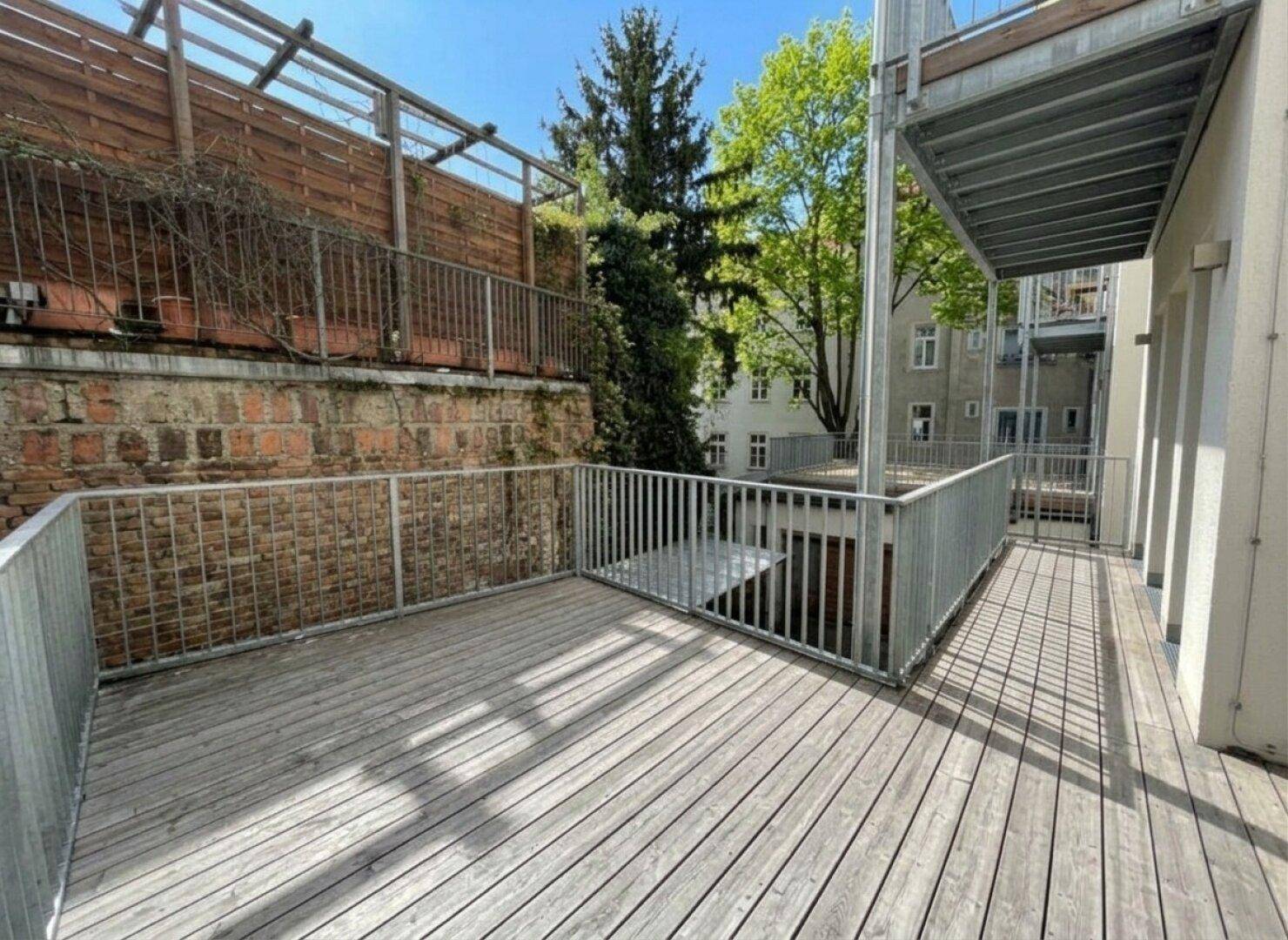 Sonnige Holzterrasse mit robustem Metallgeländer und Blick auf die umliegenden Gebäude und Bäume.