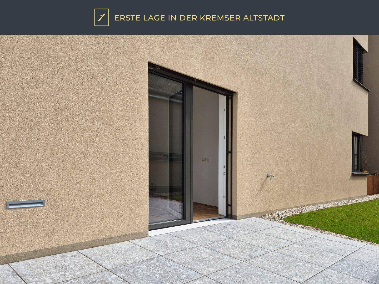 Gepflasterte Terrasse mit Rasenfläche und Zugang zu einem modernen Gebäude.