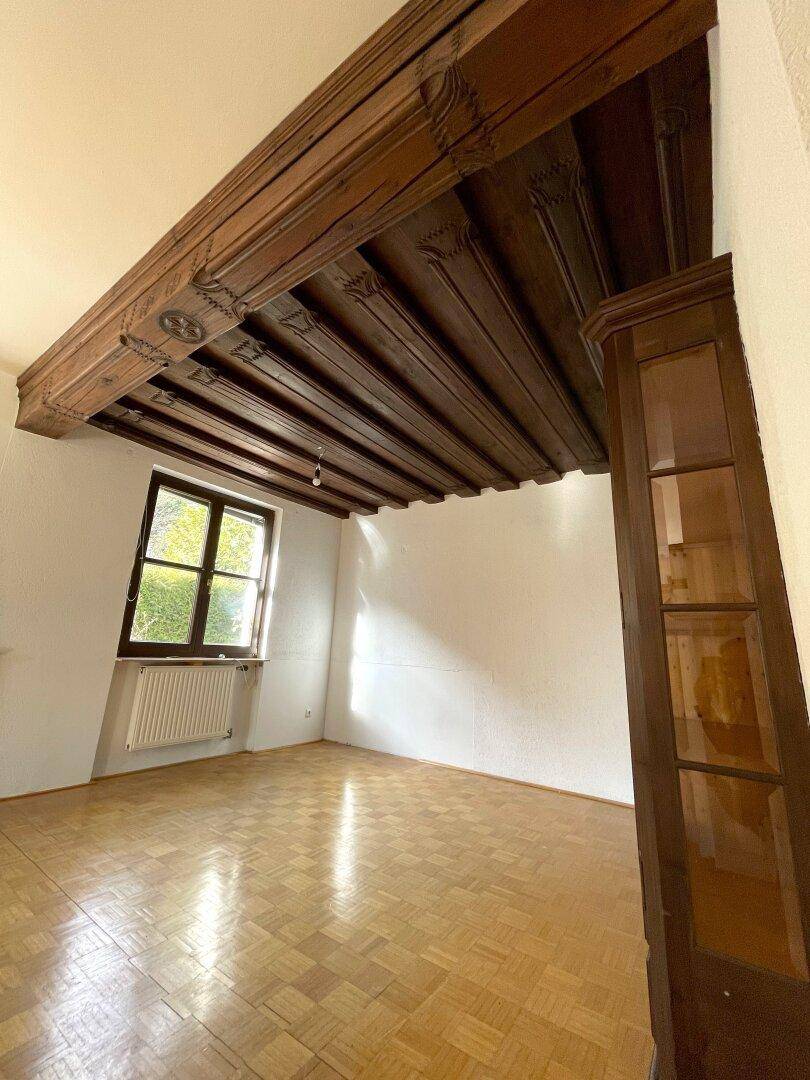 Raum mit Holzbalkendecke, Parkettboden und großem Fenster.