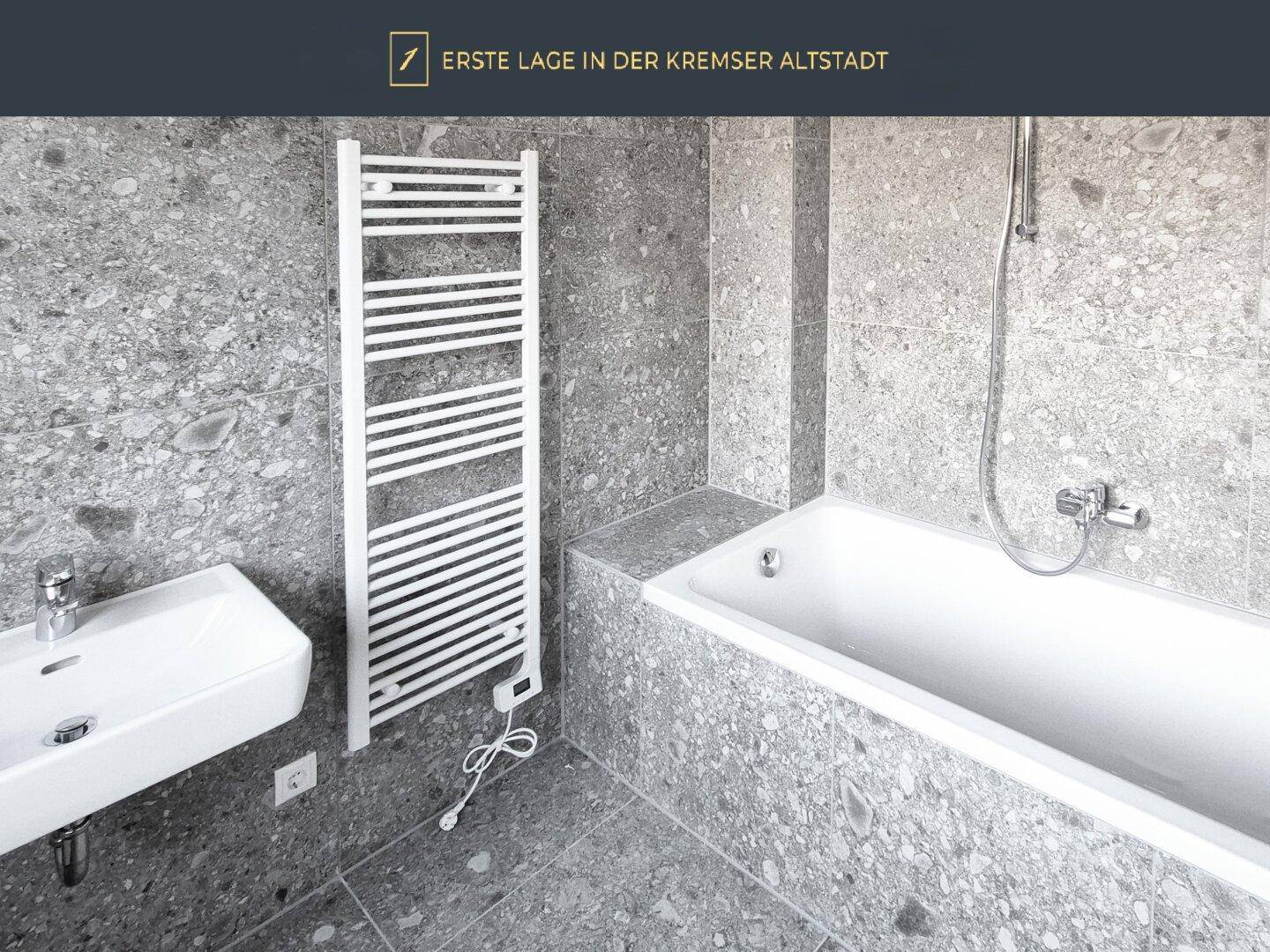Modernes Badezimmer mit Badewanne, Waschbecken, beheiztem Handtuchhalter und grauen Fliesen.