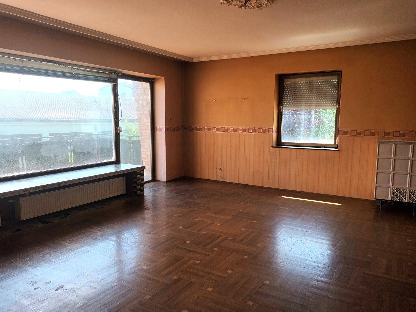 Geräumiges Wohnzimmer mit Parkettboden, großem Fenster und einem Kachelofen.