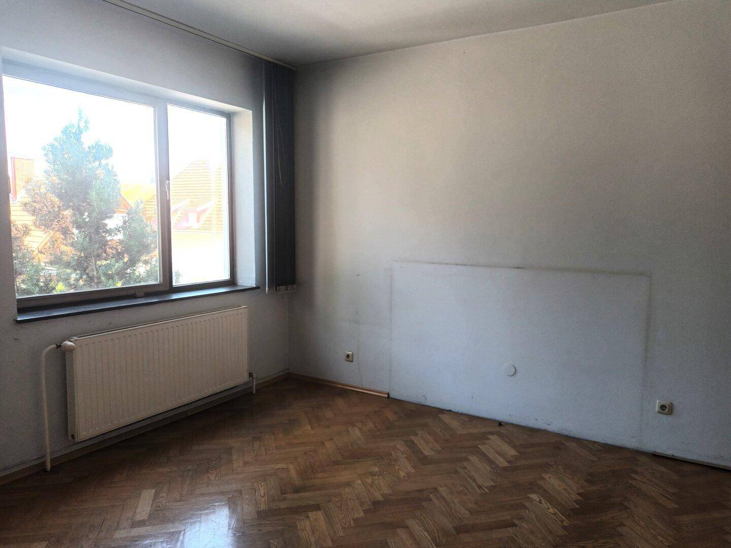 Schlafzimmer mit Parkettboden, großem Fenster und Heizkörper, bereit zur Renovierung.