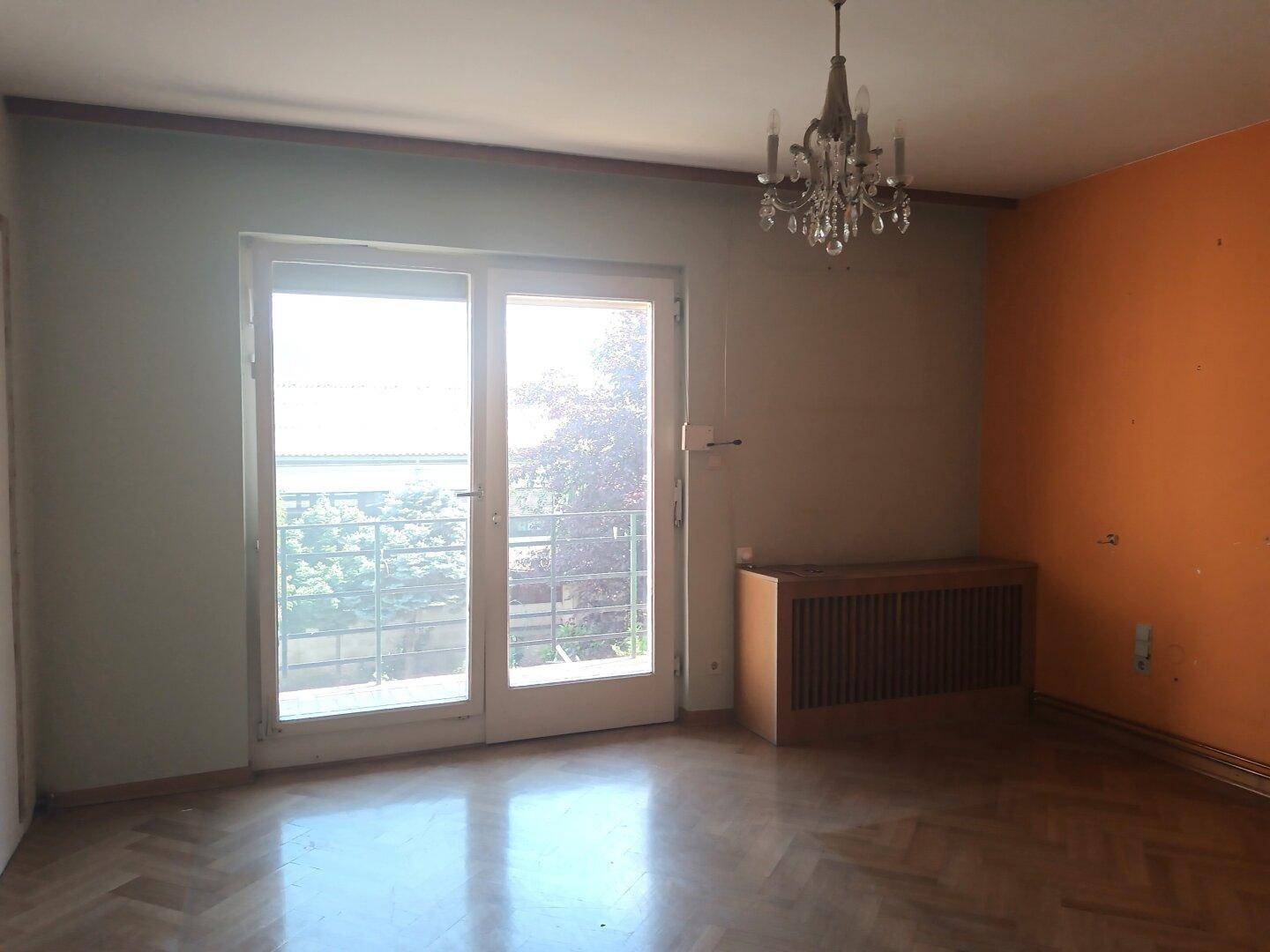 Leeres Wohnzimmer mit Parkettboden, großem Fenster und Zugang zu einem Balkon.