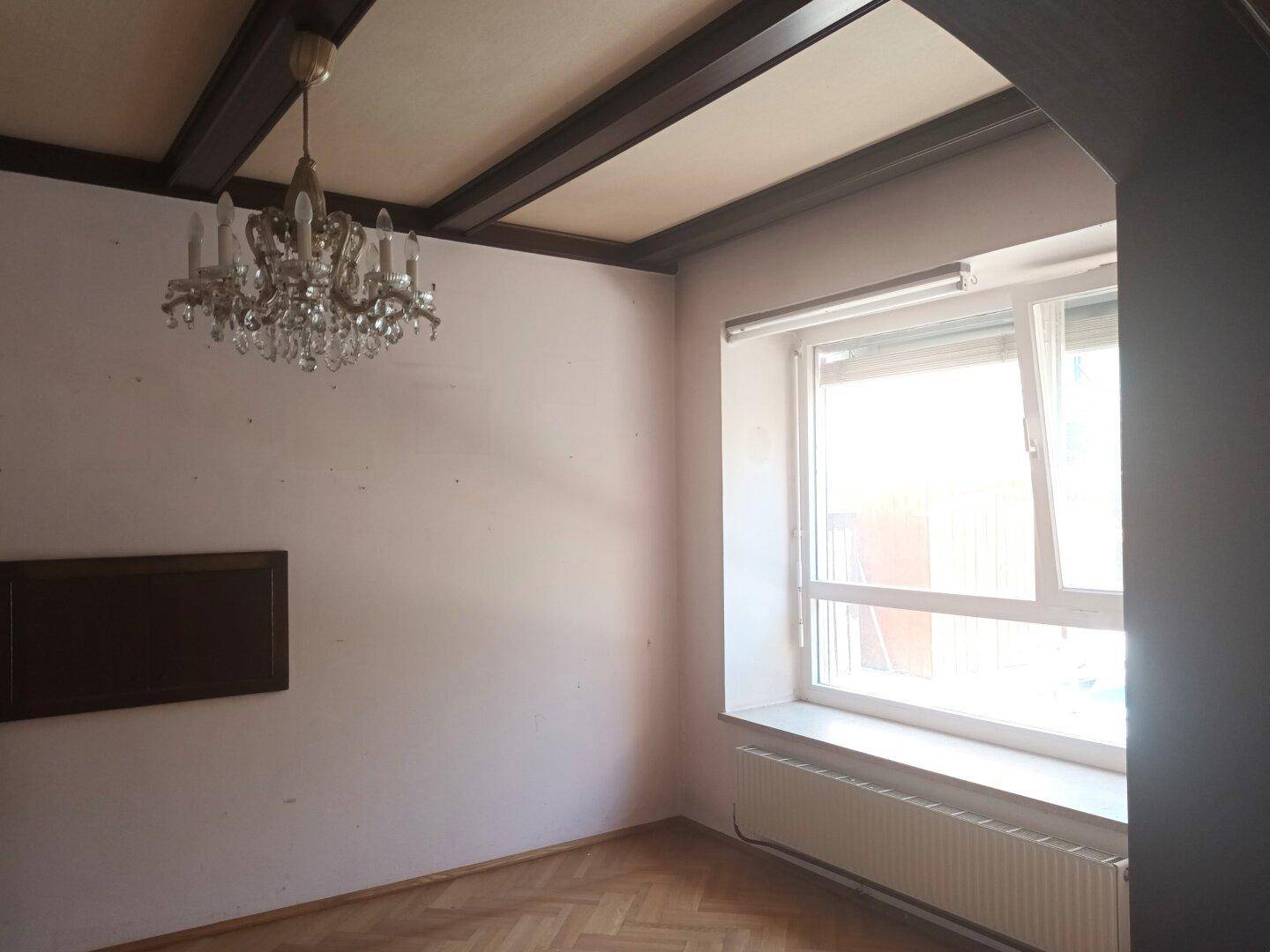 Zimmer mit Holzbalkendecke, Parkettboden, großem Fenster und Kronleuchter.