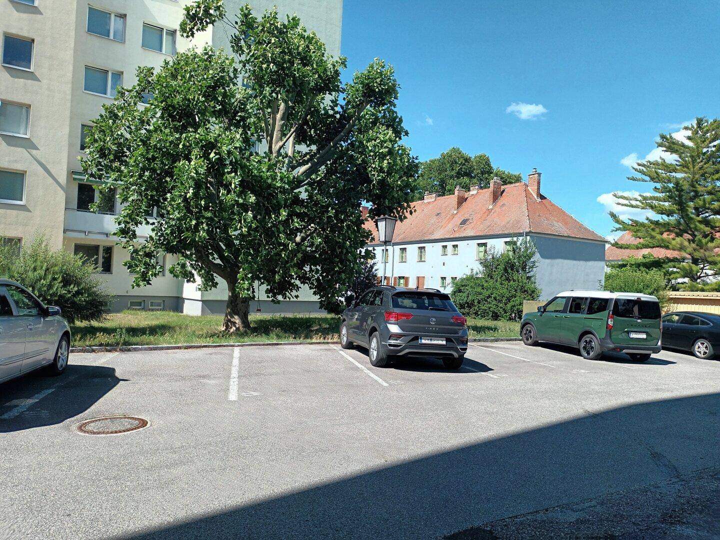 Außenansicht eines Parkplatzes mit geparkten Autos vor Wohngebäuden und Bäumen an einem sonnigen Tag.