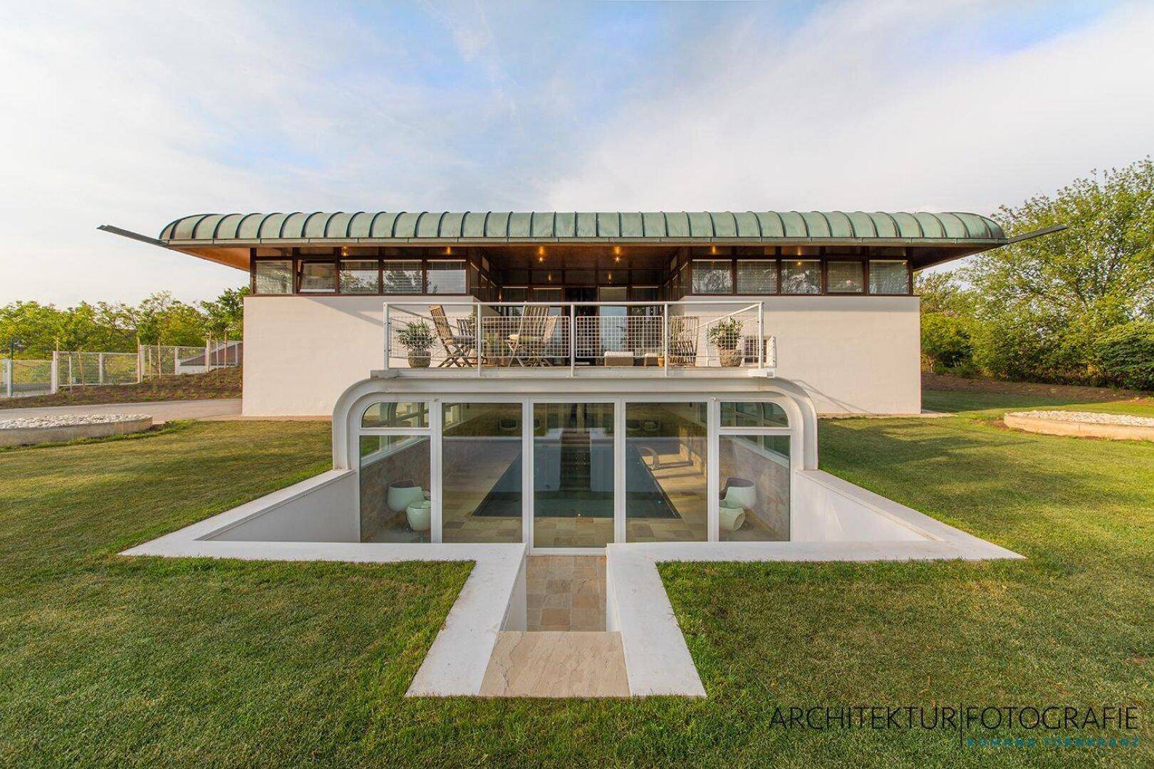 Moderne Architektur mit geschwungenem Dach, großem Garten und sichtbarem Innenpoolbereich.
