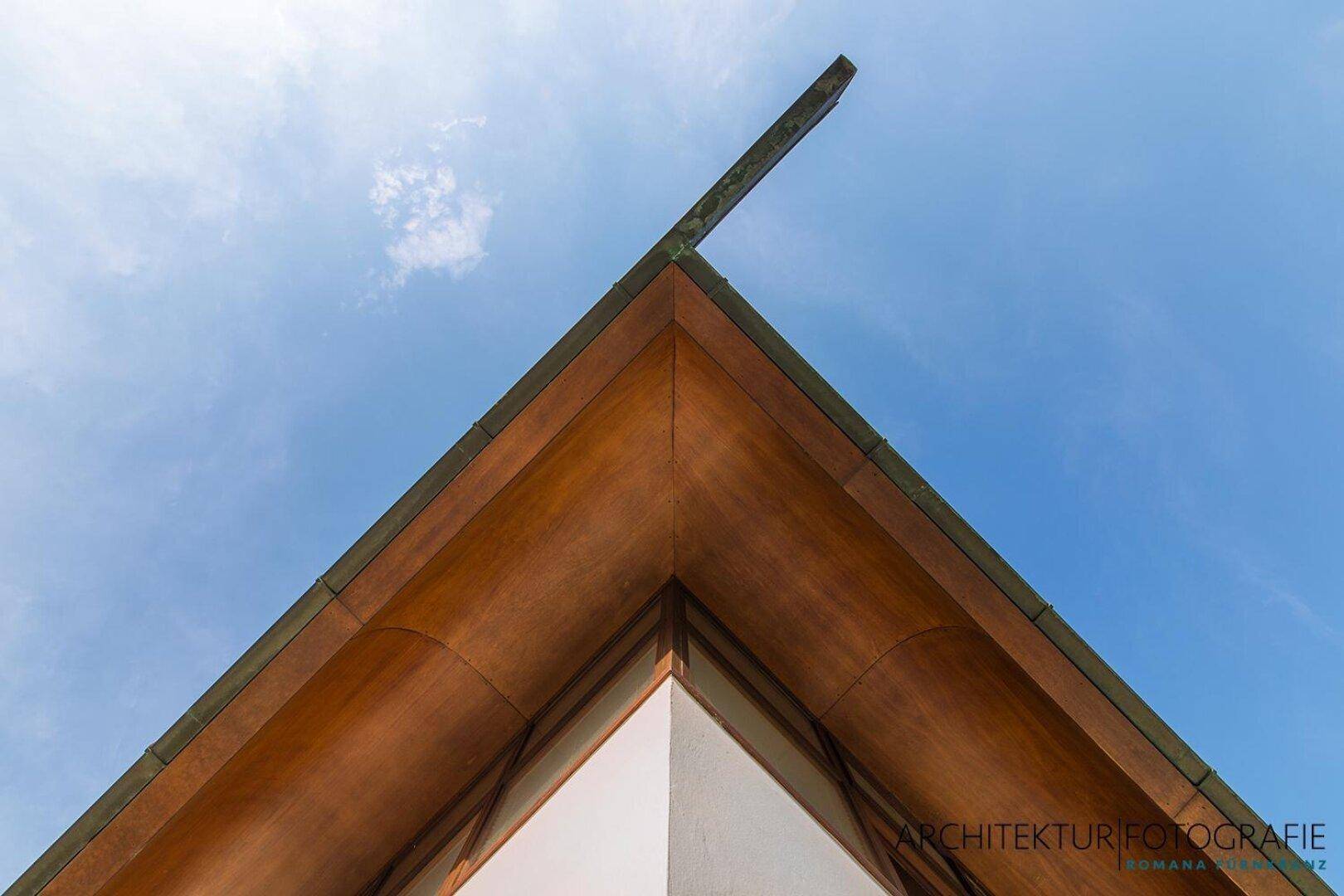 Detailaufnahme des geschwungenen Daches mit Holzuntersicht und klarem Himmel.