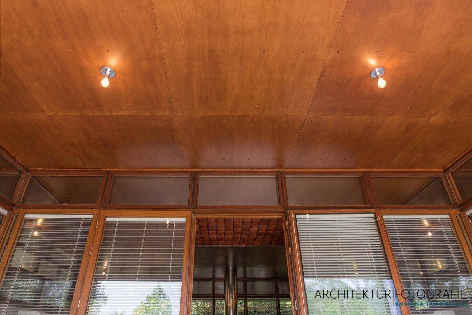 Holzverkleidete Decke über einem Bereich mit großen Fenstern und Jalousien.