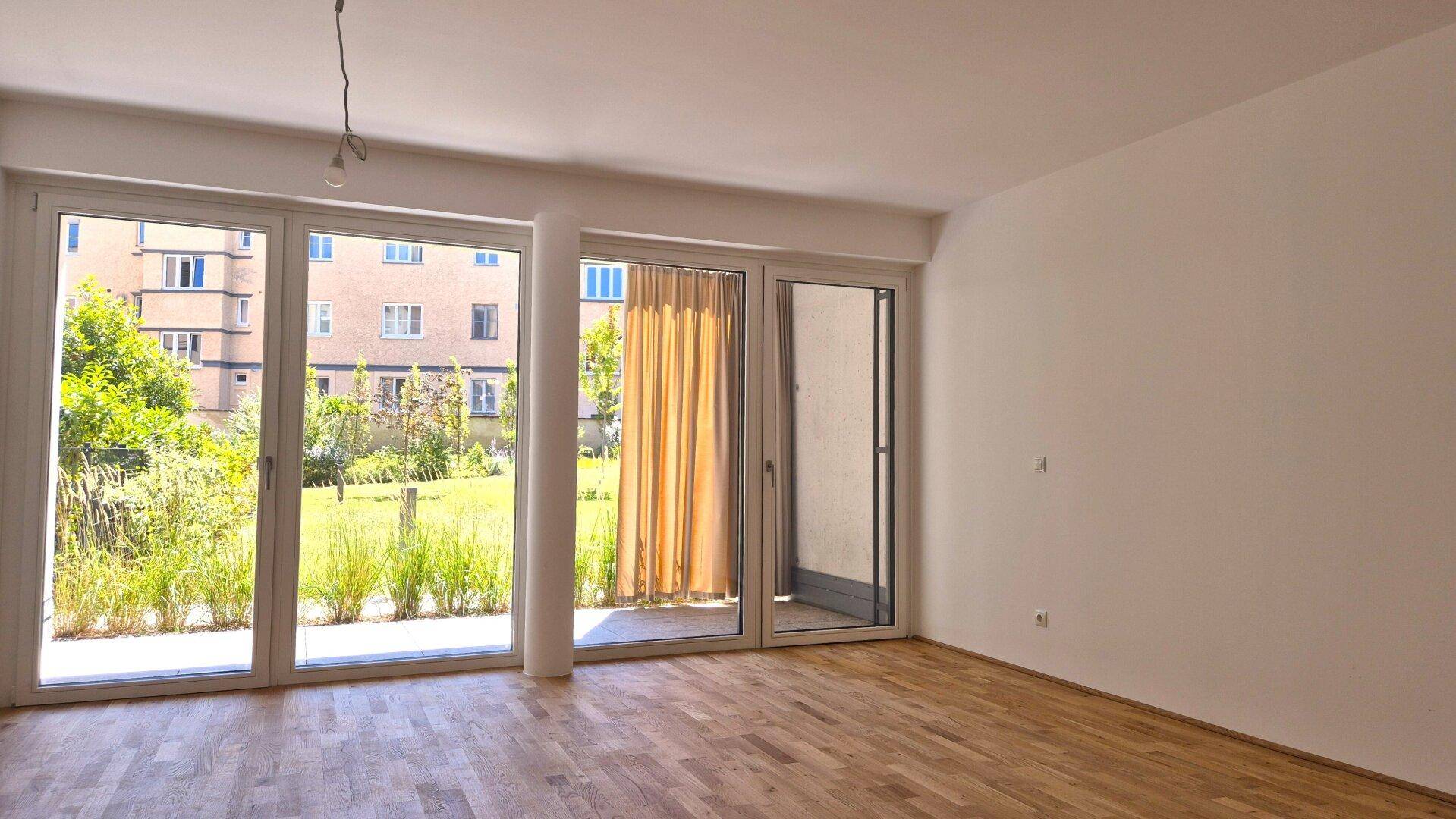 Lichtdurchfluteter Raum mit hellem Parkettboden, großen Fenstern und direktem Zugang zum Balkon mit Gartenblick.