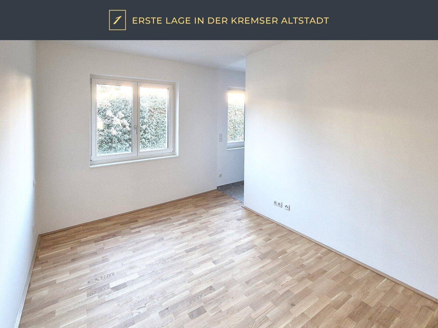 Leeres Zimmer mit Parkettboden und zwei Fenstern mit Blick ins Grüne.