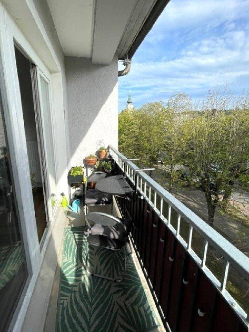 Schmaler Balkon mit Tisch und Stühlen, bietet einen Ausblick auf Bäume und einen Kirchturm.