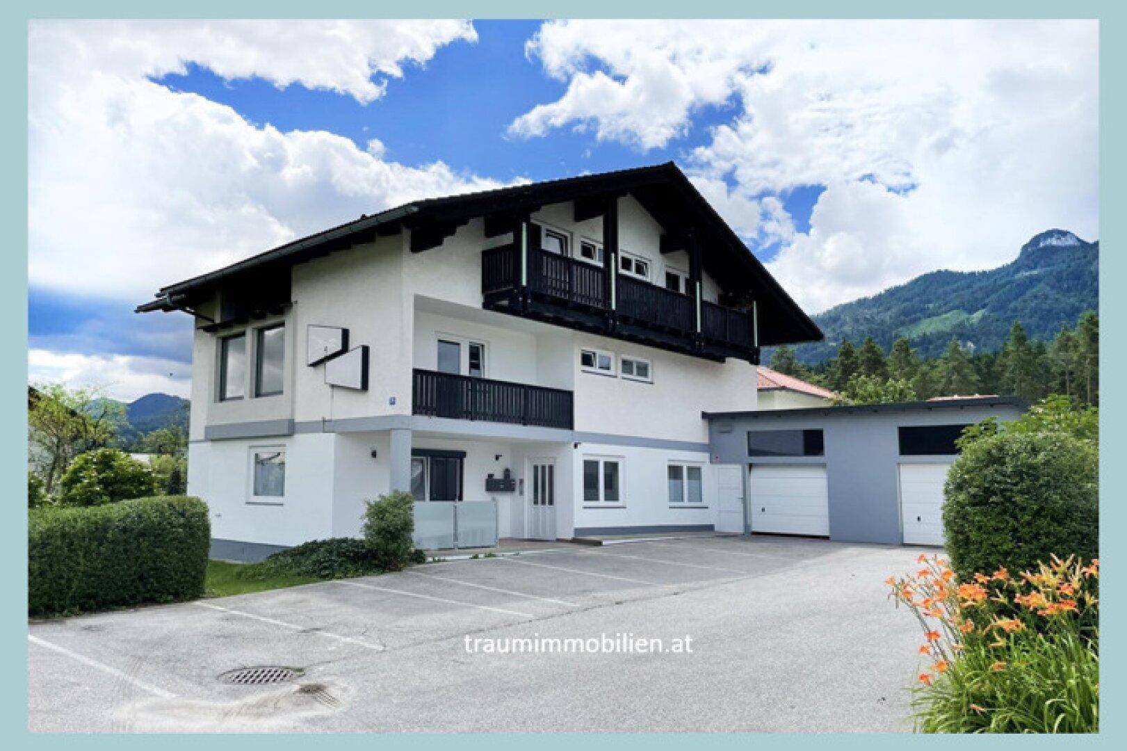 Weißes Mehrfamilienhaus mit schwarzen Balkonen, Garagen und Bergblick.