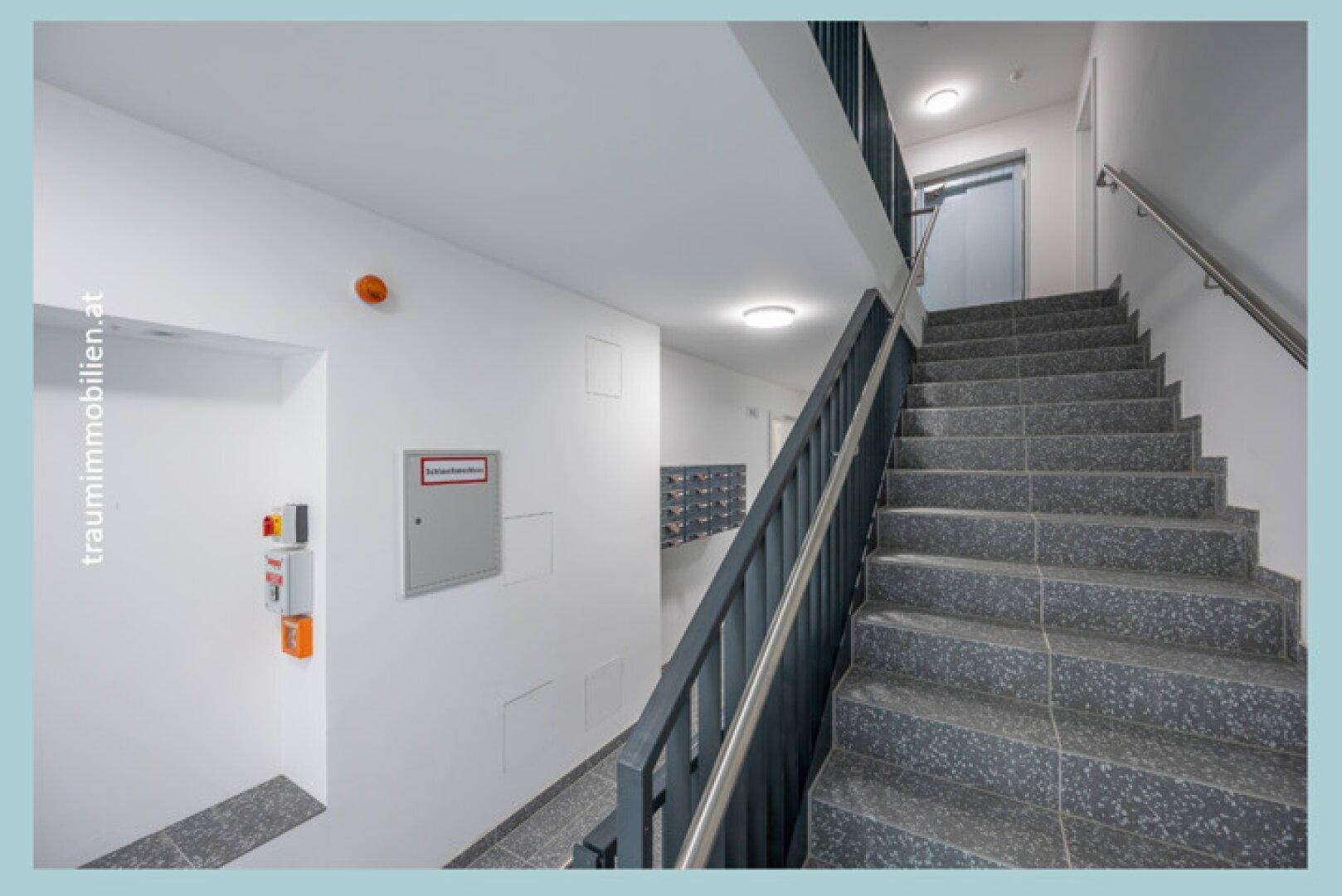 Helles Treppenhaus mit Granitstufen, Geländer und Zugang zum Aufzug.