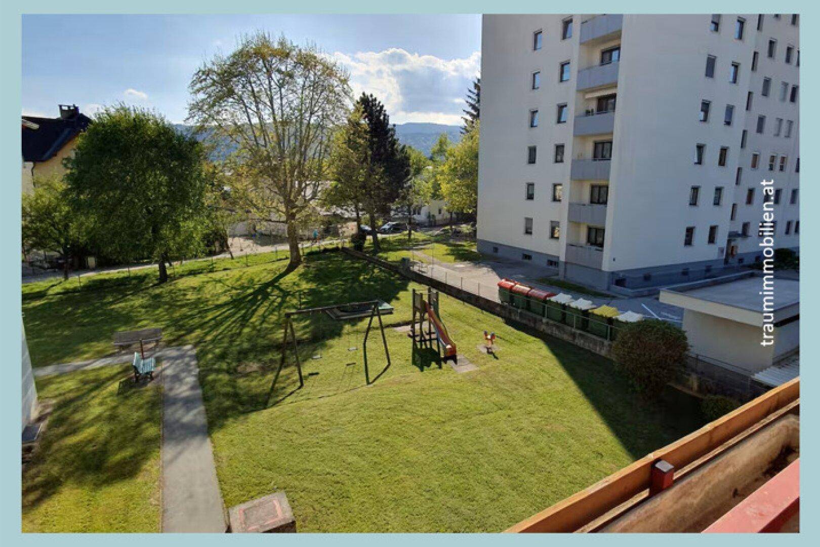 Ein gepflegter Gemeinschaftsgarten mit Spielplatz, Rasenflächen und Bäumen vor einem Wohngebäude.