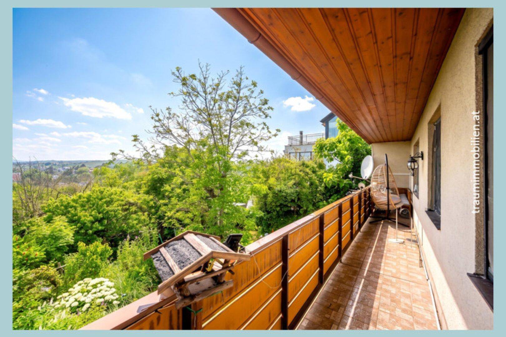 Langer Balkon mit Holzverkleidung und weitem Blick über grüne Baumwipfel und die umliegende Landschaft.