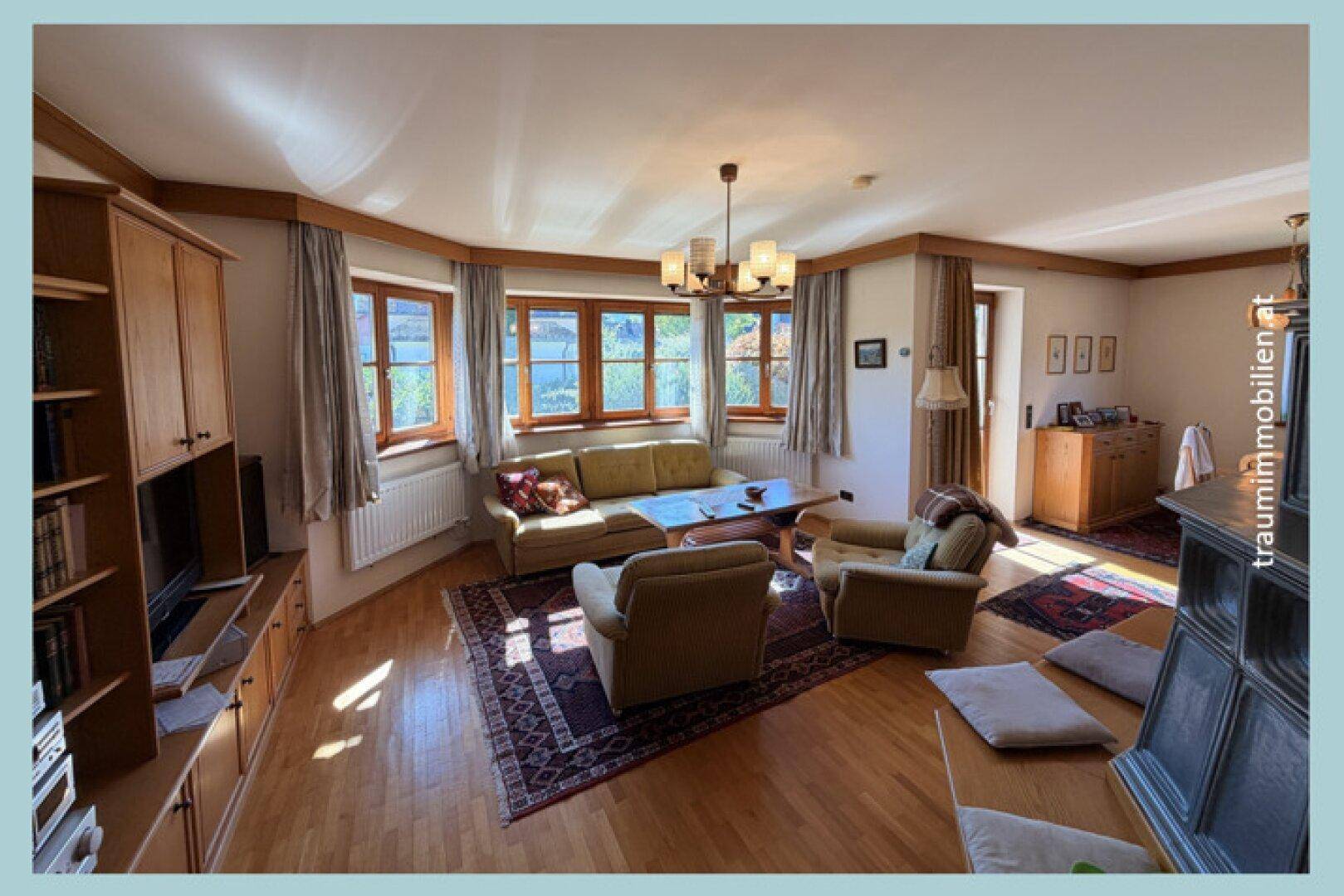 Gemütliches Wohnzimmer mit Holzboden, großen Fenstern und Kachelofen.