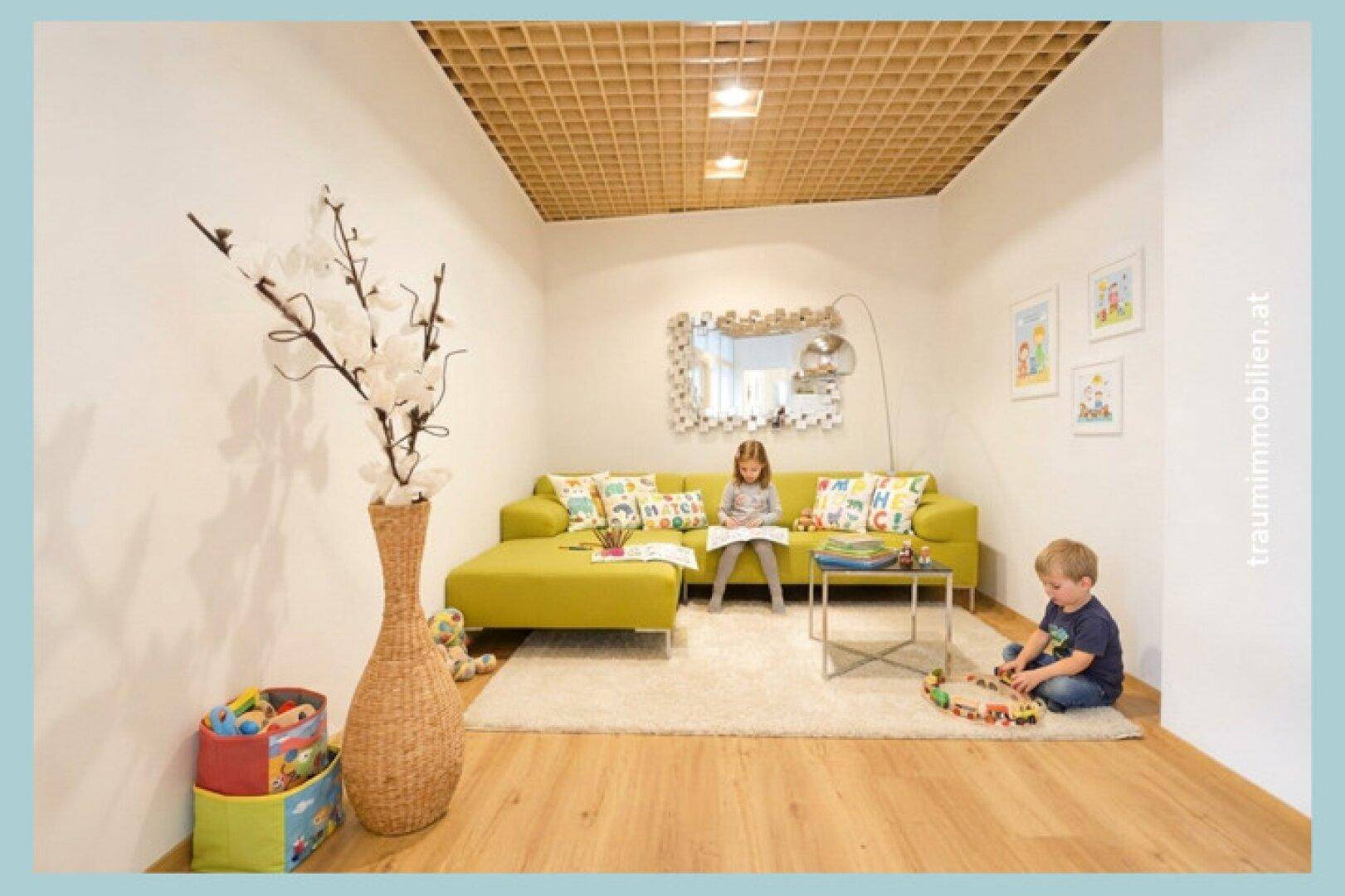 Kinderfreundlicher Wartebereich mit grüner Couch, Teppich und Holzlamellendecke, ideal zum Spielen.