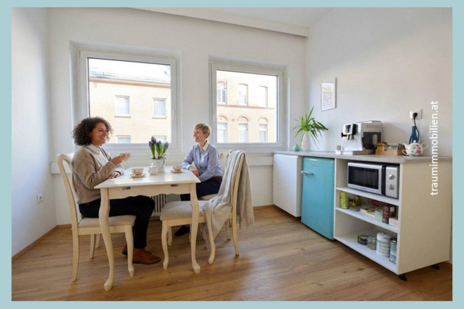 Heller Pausenraum mit Esstisch, Stühlen, Fenstern und einer kleinen Küchenzeile mit Kühlschrank und Mikrowelle.