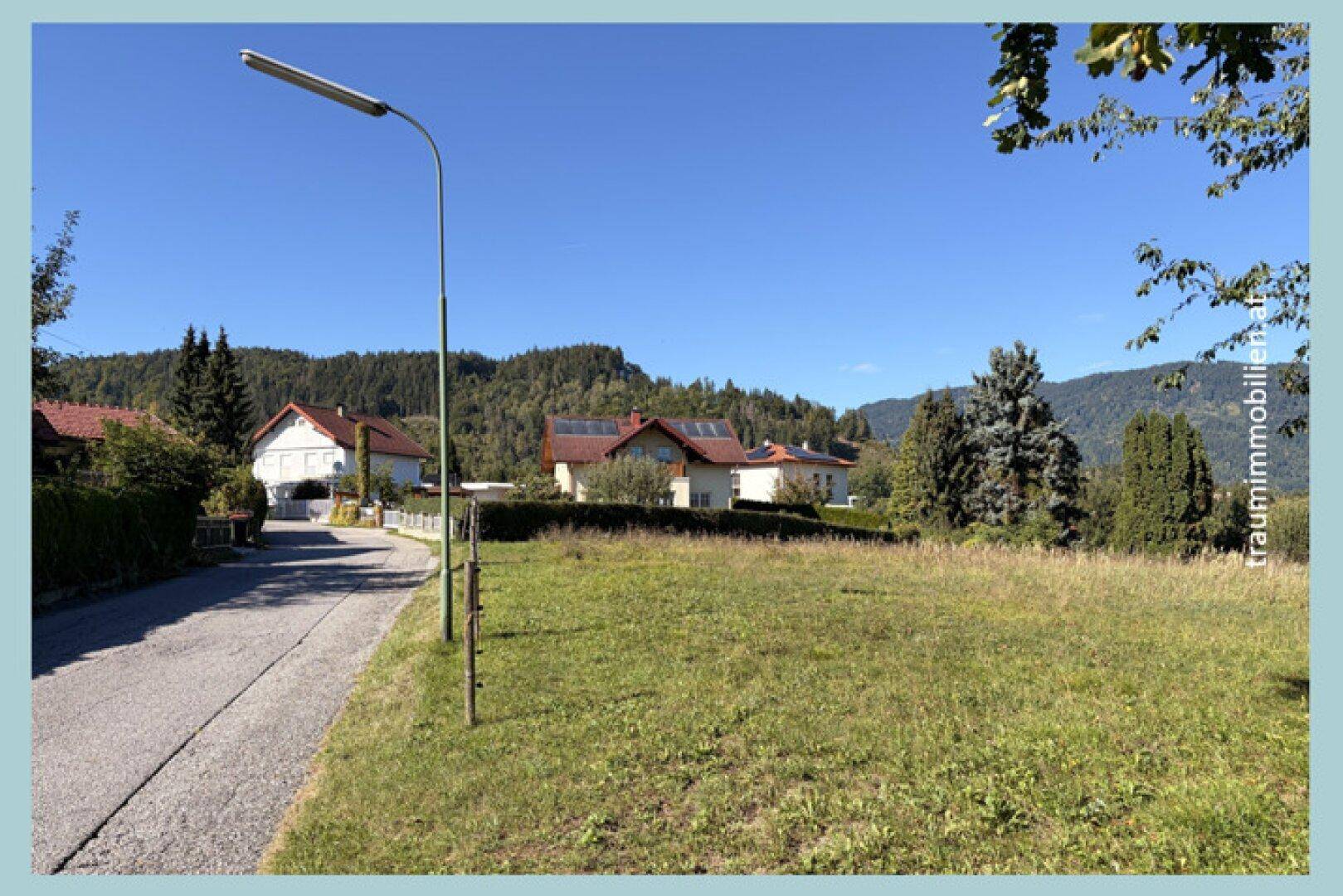 Weitläufiges Baugrundstück in ländlicher Umgebung mit Blick auf umliegende Häuser und Berge unter blauem Himmel.