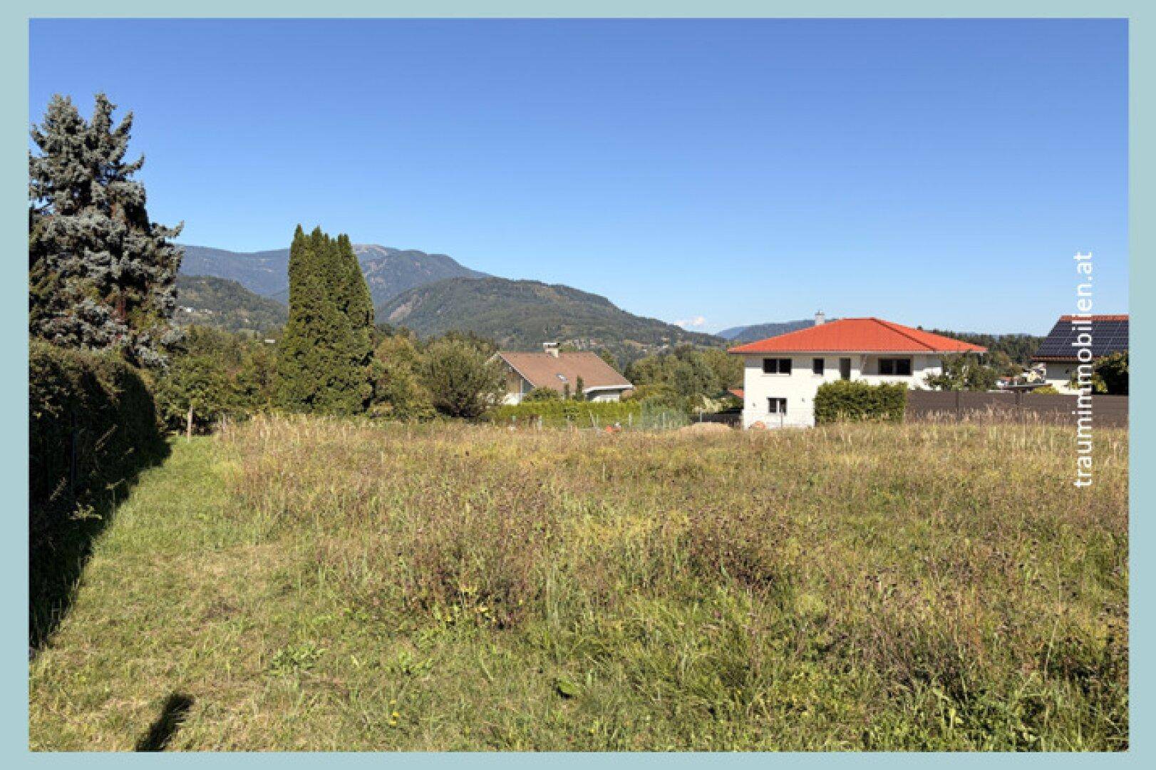 Grüne Wiese als Baugrundstück mit angrenzenden Wohnhäusern und einer malerischen Bergkulisse im Hintergrund.
