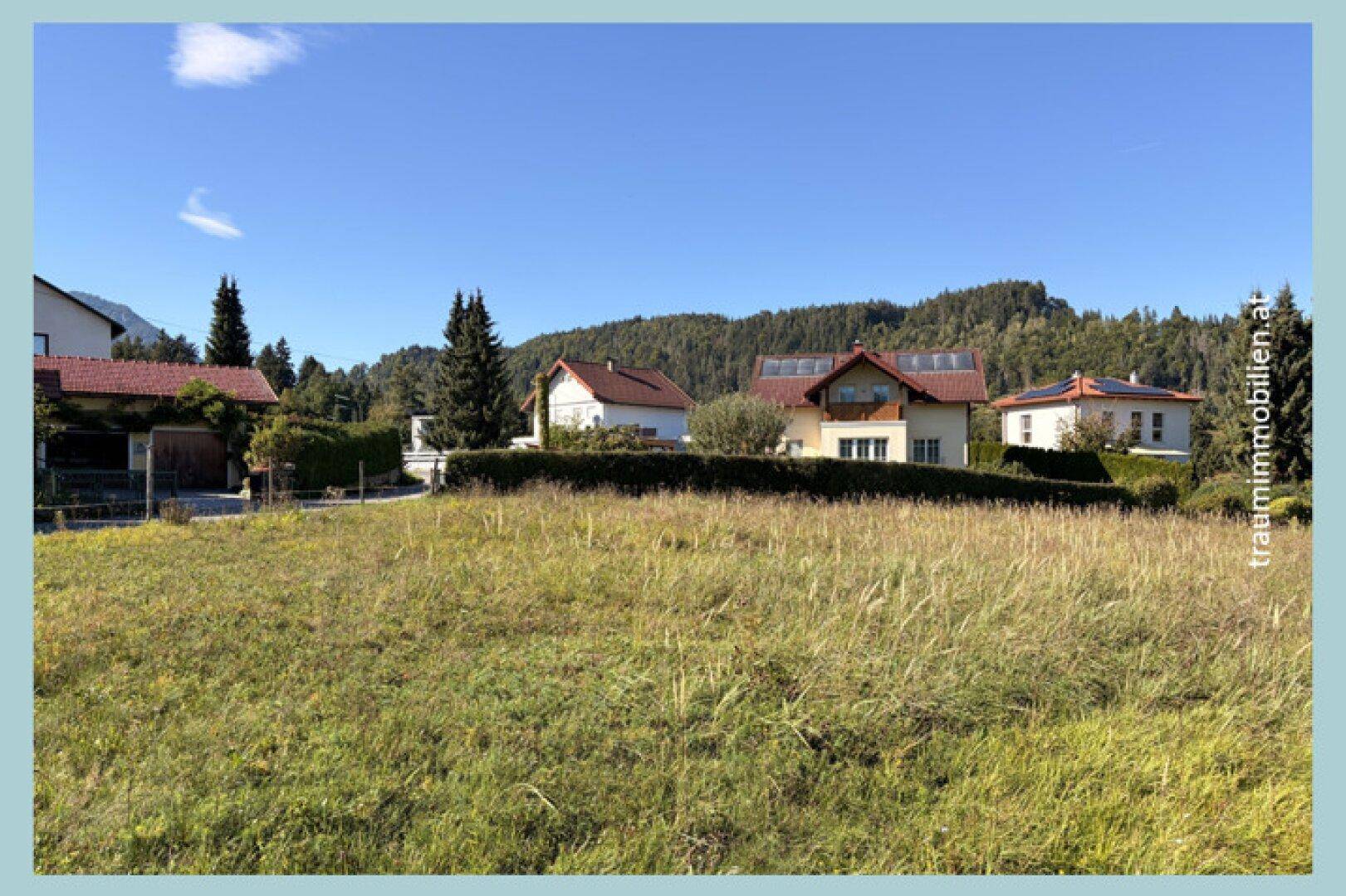 Großes Wiesengrundstück mit Blick auf moderne Wohnhäuser, einige mit Solaranlagen, und die Bergkette.