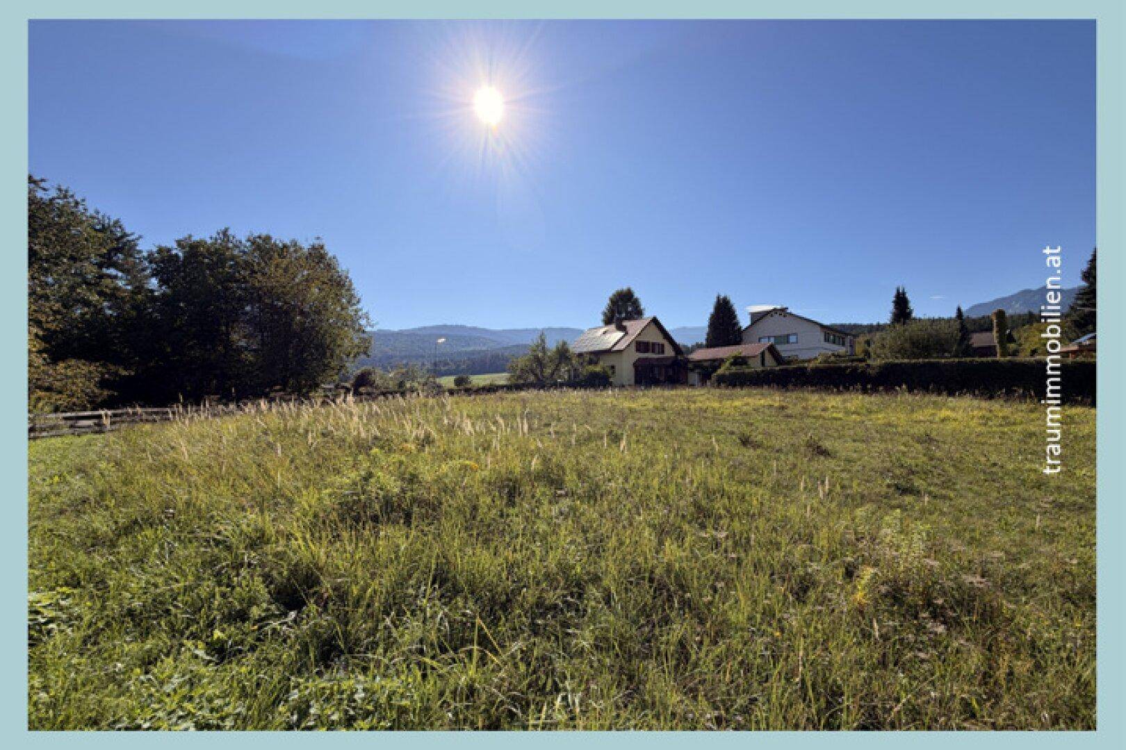 Helles Wiesengrundstück unter strahlendem Sonnenschein, umgeben von Häusern und einer beeindruckenden Berglandschaft.
