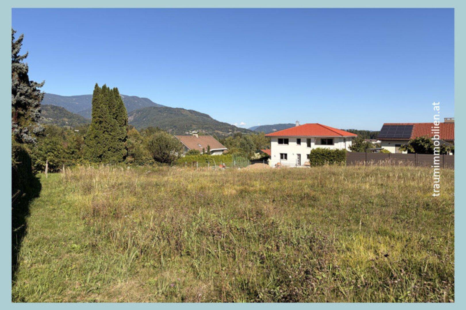 Großes Wiesengrundstück mit Blick auf Wohnhäuser und die beeindruckende Bergkulisse unter blauem Himmel.
