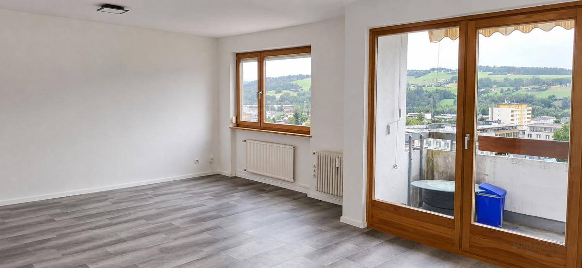 Lichtdurchfluteter Wohnraum mit grauem Laminatboden und weitem Blick auf die grüne Umgebung.