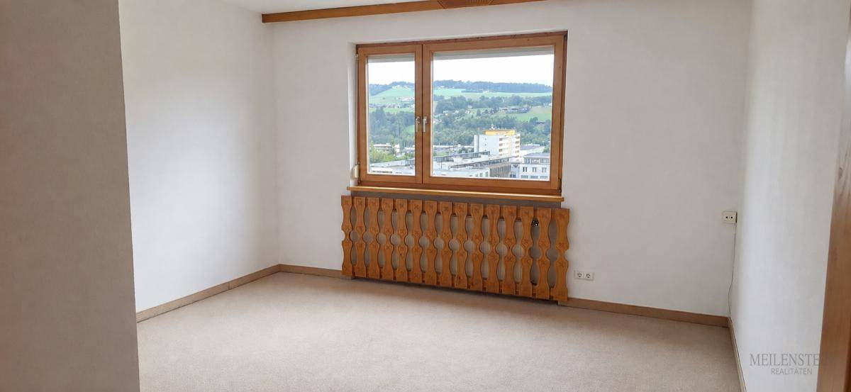 Helles Zimmer mit Teppichboden und einem Fenster, das einen weiten Blick ins Grüne bietet.