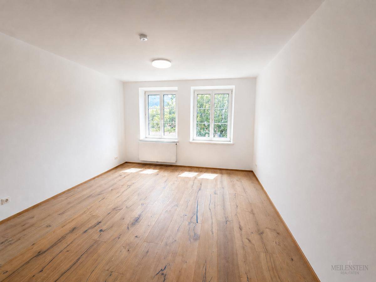 Leeres Zimmer mit hellem Holzboden und zwei Fenstern, die einen Blick ins Grüne bieten.