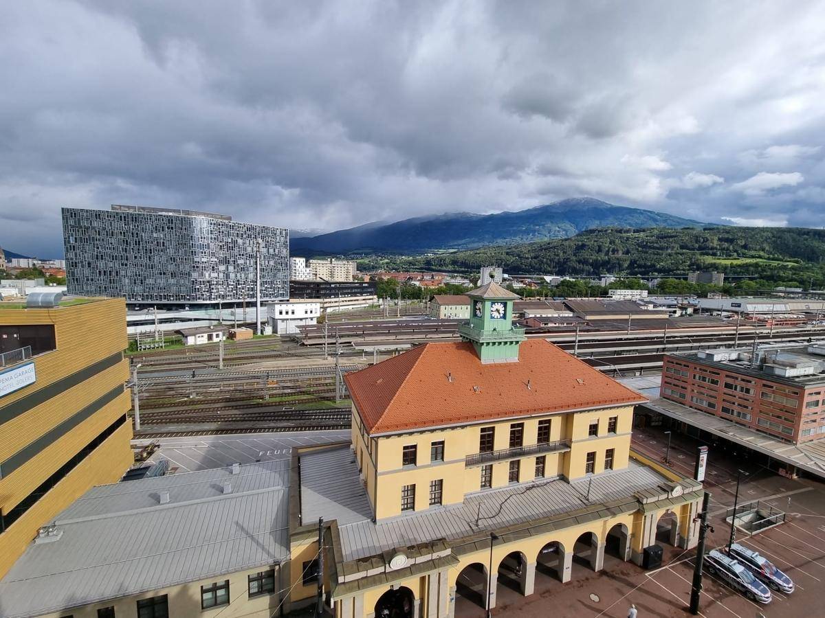 Panoramablick auf die Stadt mit Bahnhof, umliegenden Gebäuden und majestätischen Bergen im Hintergrund.