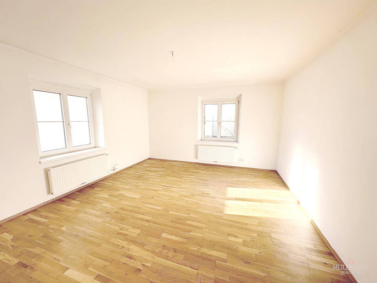 Helles, leeres Zimmer mit Parkettboden und zwei Fenstern, das viel Tageslicht hereinlässt.