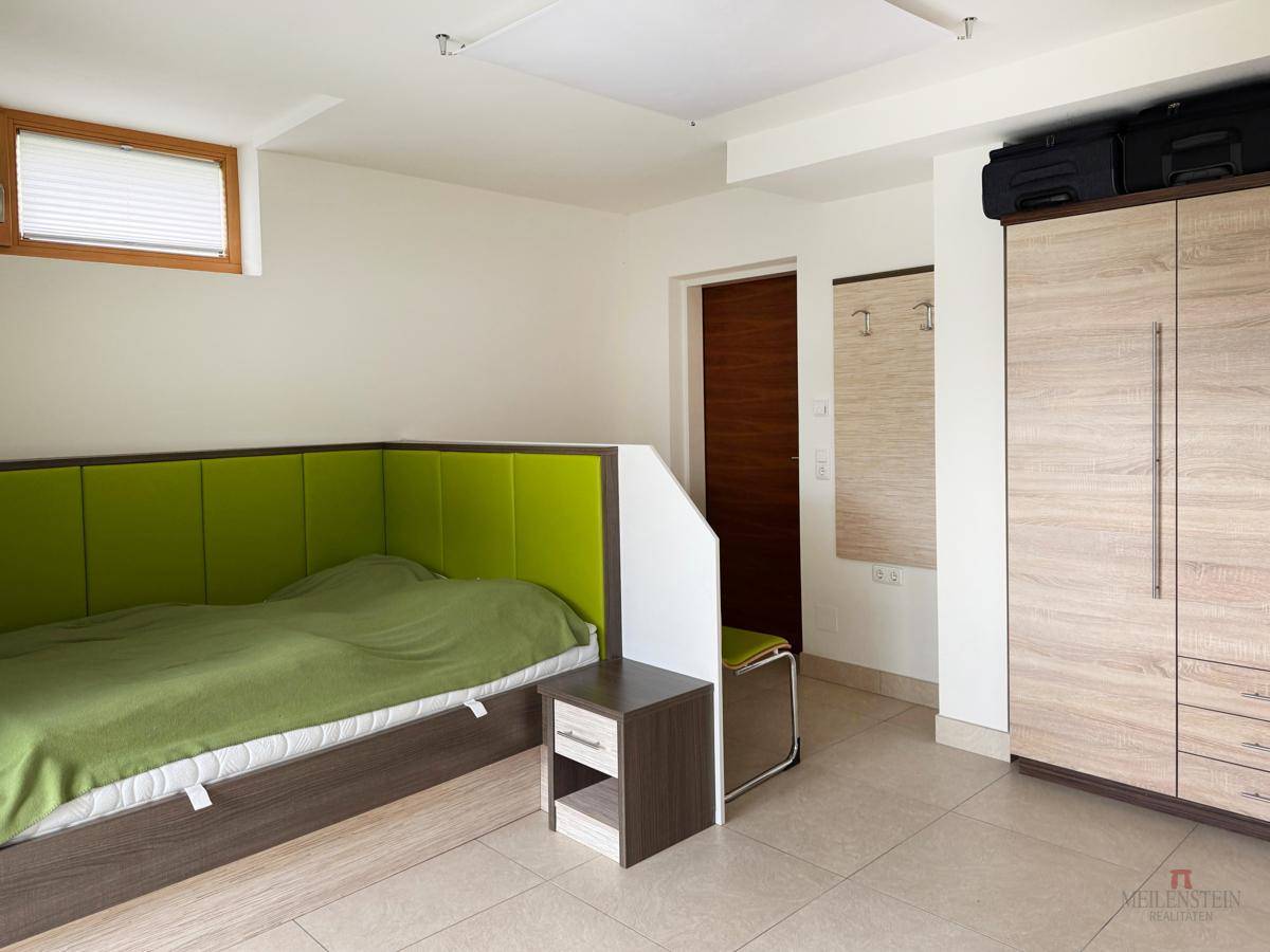 Helles Schlafzimmer mit grünem Kopfteil, Kleiderschrank und Fenster, einfach eingerichtet.