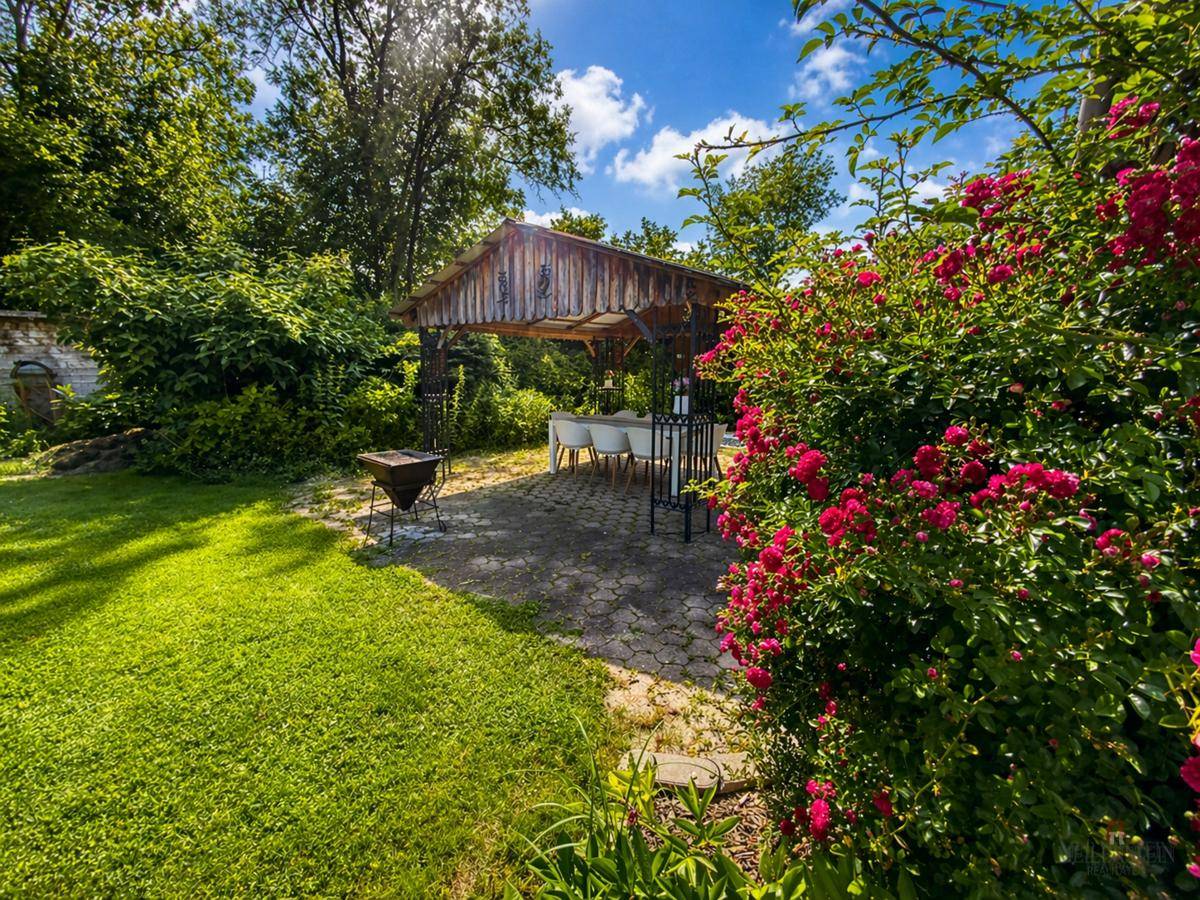 Idyllischer Garten mit überdachtem Sitzbereich, Grillplatz und blühenden Rosenbüschen.