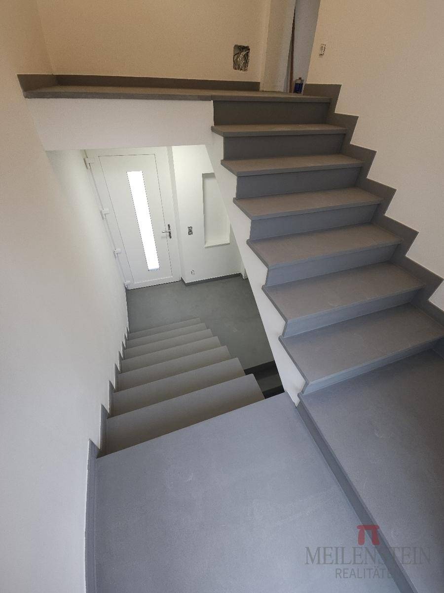 Moderne Treppe mit grauen Stufen und weiß gestrichenen Wänden, führt in die unteren Etagen.