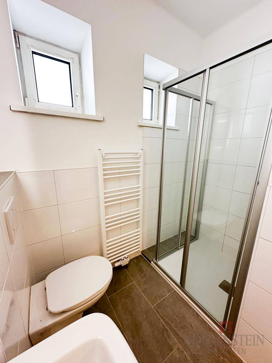 Duschbereich im Badezimmer mit Glasabtrennung, WC und Fenster, hell gefliest.