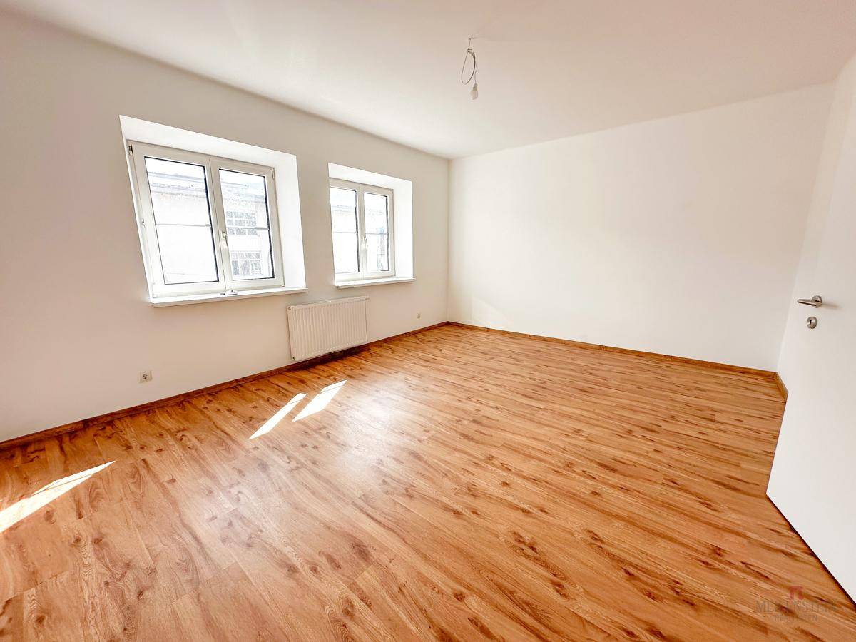 Geräumiges, helles Zimmer mit Holzboden und weiß gestrichenen Wänden, inklusive Türzugang.