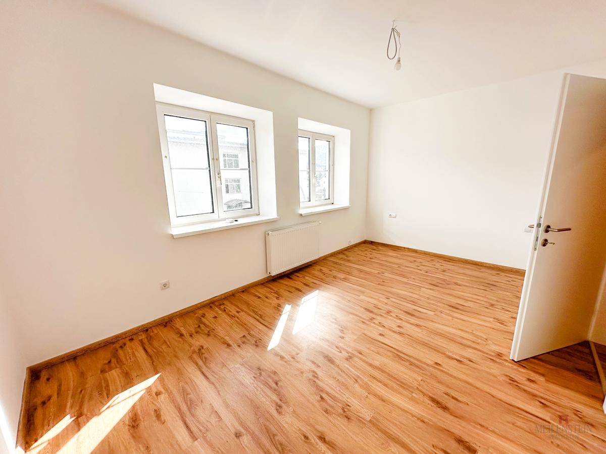 Lichtdurchflutetes Zimmer mit Holzboden und zwei Fenstern, bietet viel Platz für individuelle Einrichtung.