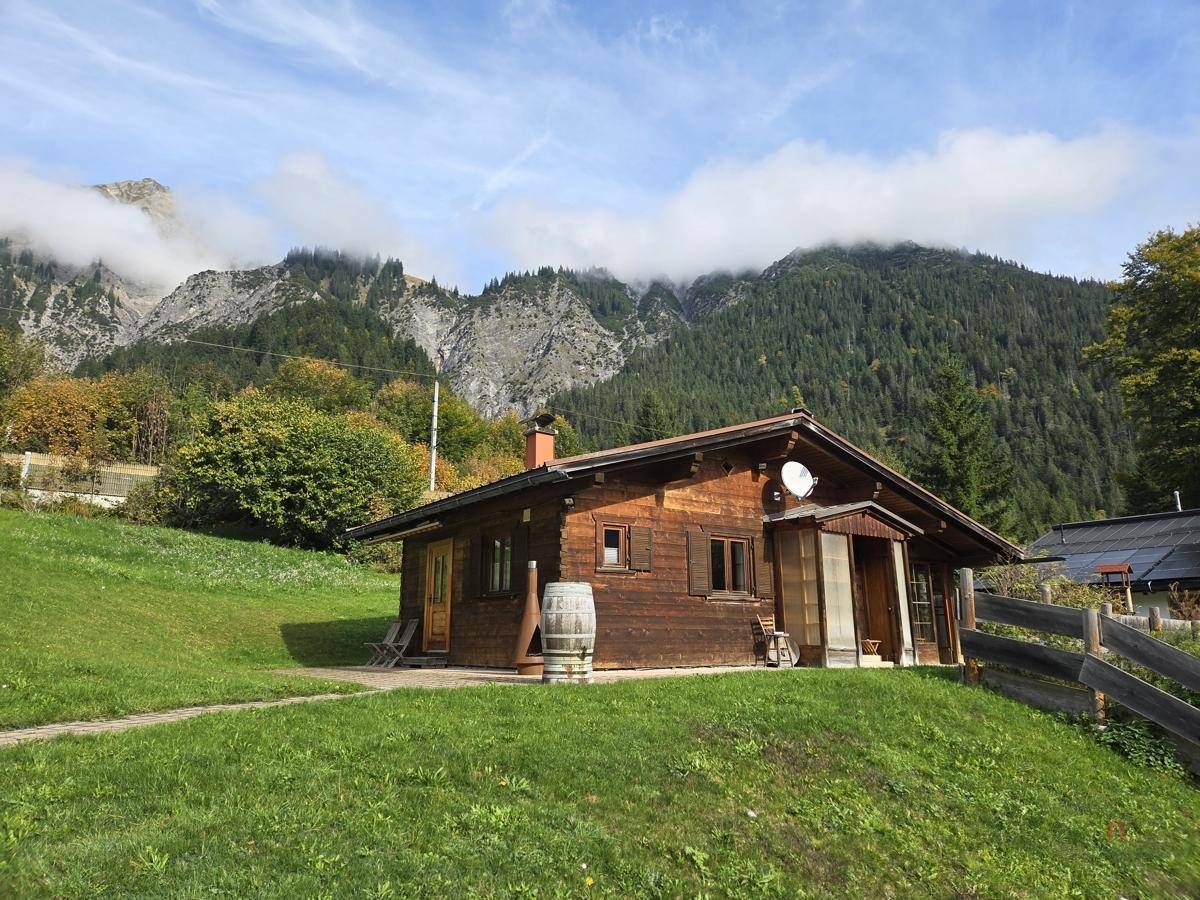 Rustikales Holzhaus mit grüner Wiese und bewaldeten Bergen im Hintergrund unter bewölktem Himmel.