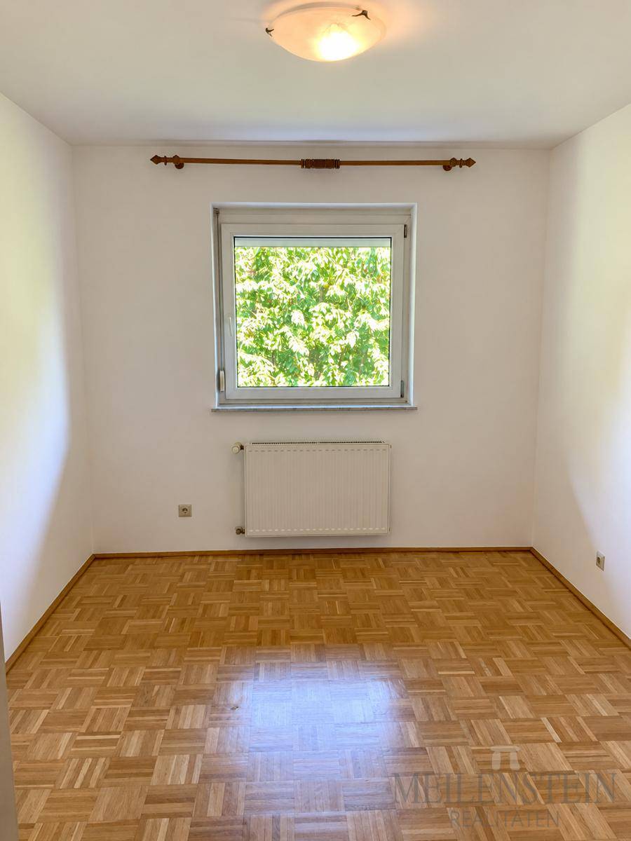 Leeres Zimmer mit Parkettboden und Fenster, das einen Blick ins Grüne bietet.