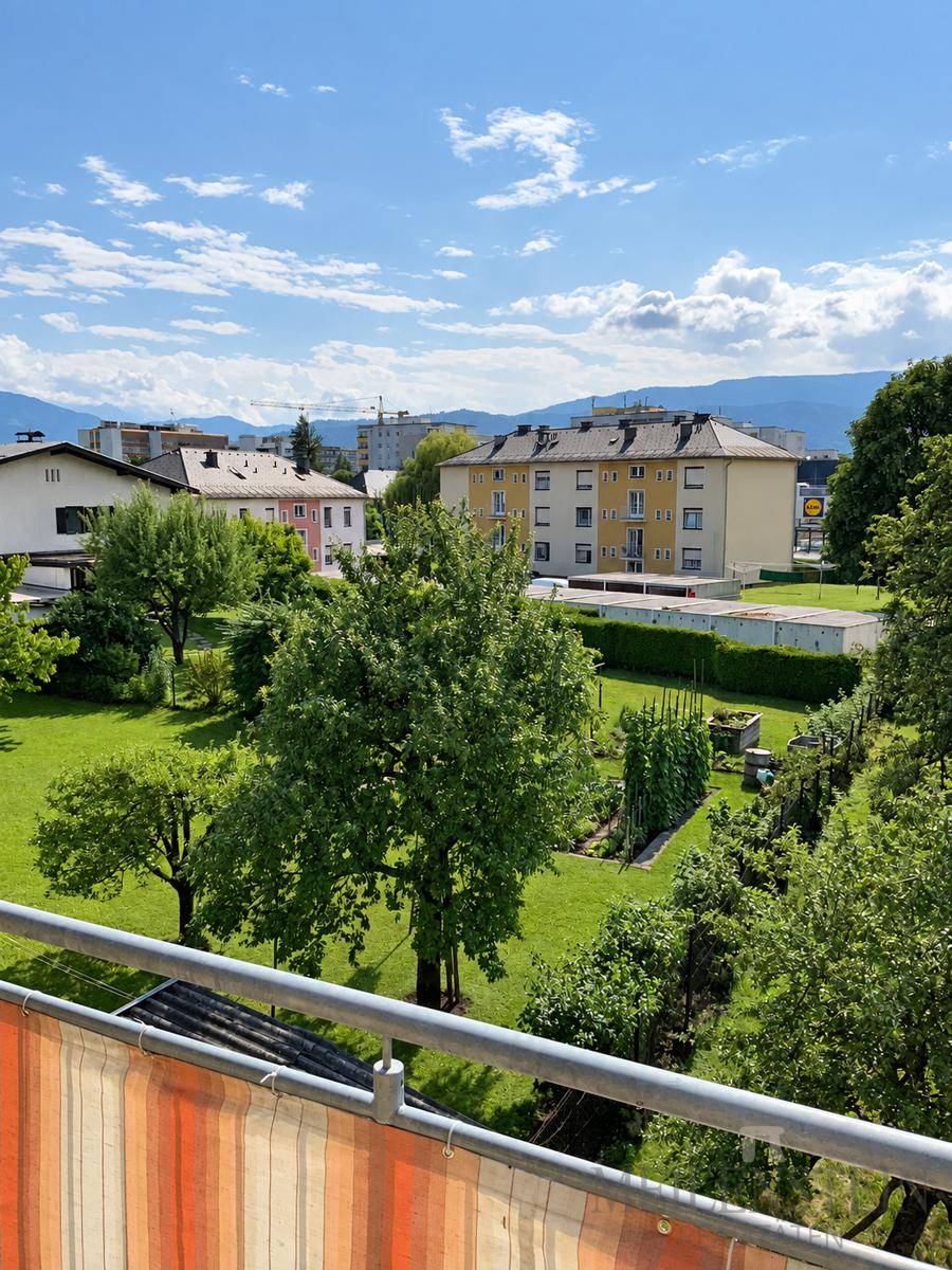 Panoramablick vom Balkon auf weitläufige Grünflächen, Wohngebäude und ferne Berge.