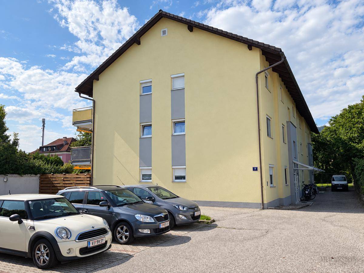 Ansicht des Mehrfamilienhauses mit gelber Fassade und Parkplätzen vor dem Gebäude.