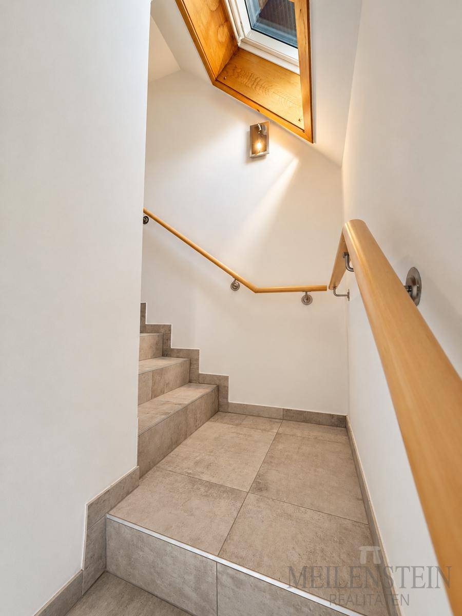 Treppenaufgang mit Holzgeländer, Fliesenboden und einem Dachfenster für natürliches Licht.