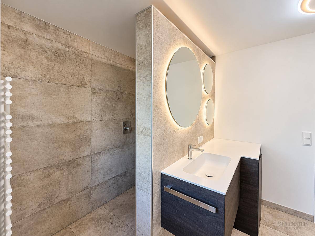 Modernes Badezimmer mit begehbarer Dusche, beleuchtetem Spiegel und Einzelwaschbecken.