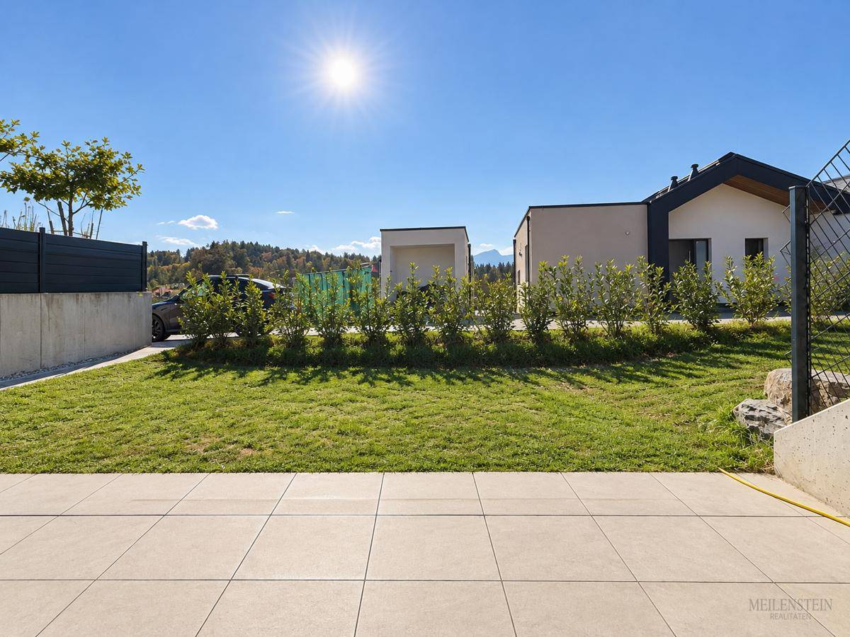 Sonniger Gartenbereich mit Rasenfläche und gepflasterter Terrasse, umgeben von Hecken.
