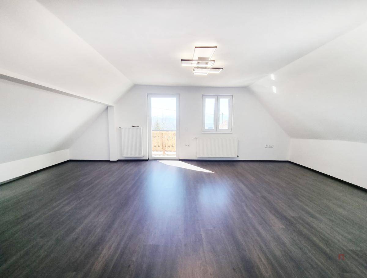Helles, leeres Zimmer im Dachgeschoss mit dunklem Laminatboden und Zugang zu einem kleinen Balkon.